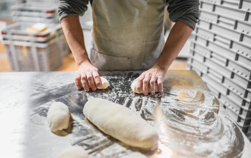 Panadeiro preparando a masa para cocer pan e pastelería, traballando con fariña sobre unha mesa de aceiro inoxidable nunha cociña profesional de panadaría.
